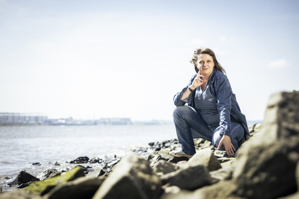 Die Kursleitende für Zeichnen der Volkshochschule Anna Goldmund hockt mit wehendem Haar und Mantel an der Steinkante des Elbstrandes in Blankenese